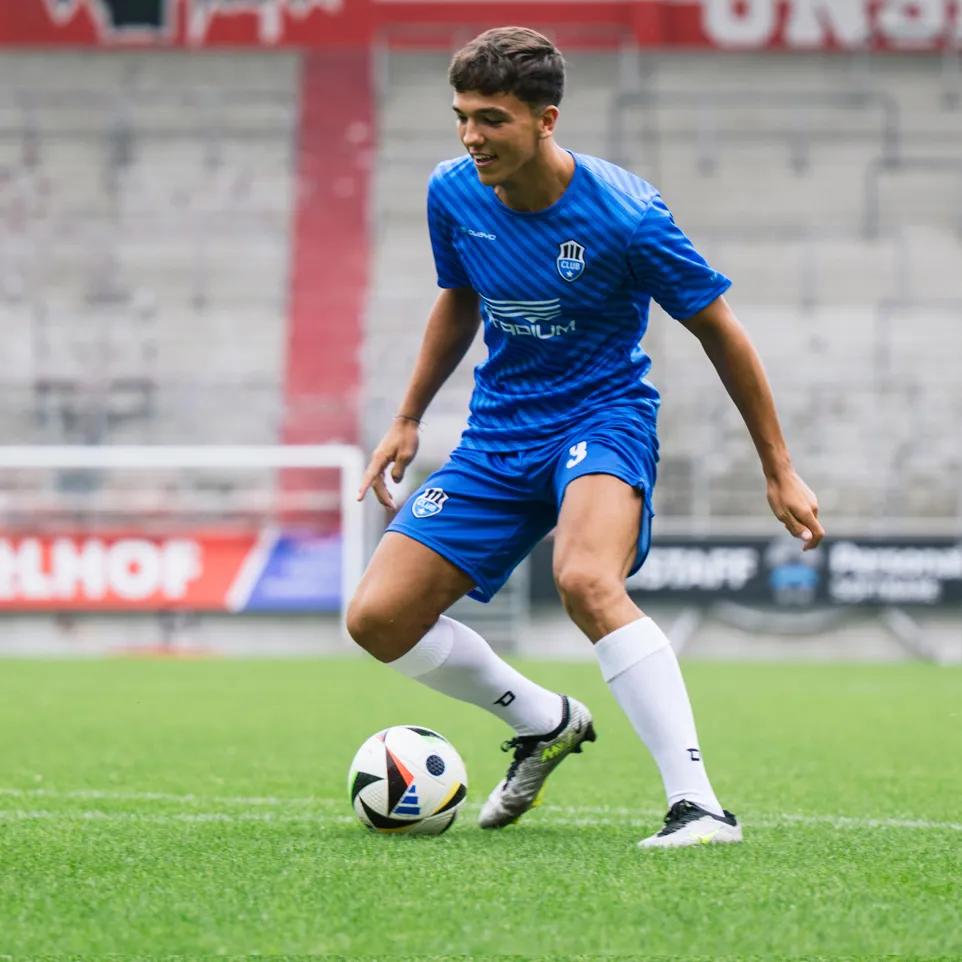 Un joven futbolista con una camiseta azul y pantalones cortos azules con un logo en el pecho y el número 3 en los pantalones cortos controla un balón de fútbol en un campo de césped verde.